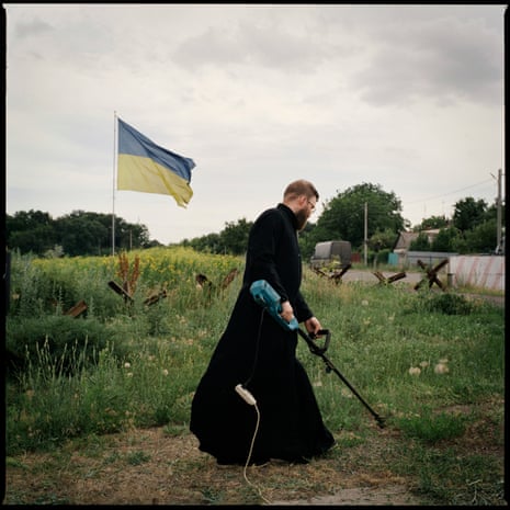 A man in a black clergy dress strims the grass next to a Ukrainian flag