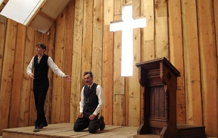 Paul Dano points at Daniel Day-Lewis, kneeling, on a stage with a cross behind them in There Will Be Blood