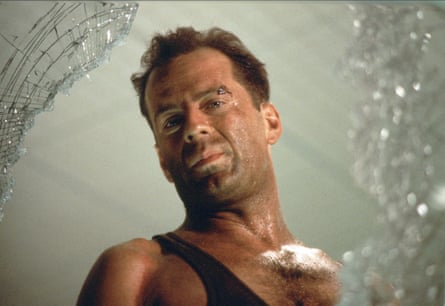 Bruce Willis as John McClane in Die Hard, wearing a black vest and looking through a broken glass window