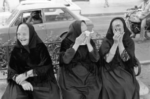 Three older women in dark dresses and headscarves sitting on a bench, smiling or laughing. Two are covering their faces