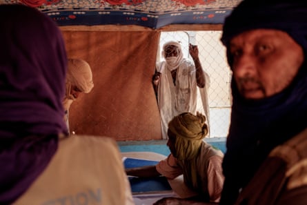 Men with scarves on their heads and faces in a tent, with a man looking in from behind a net.