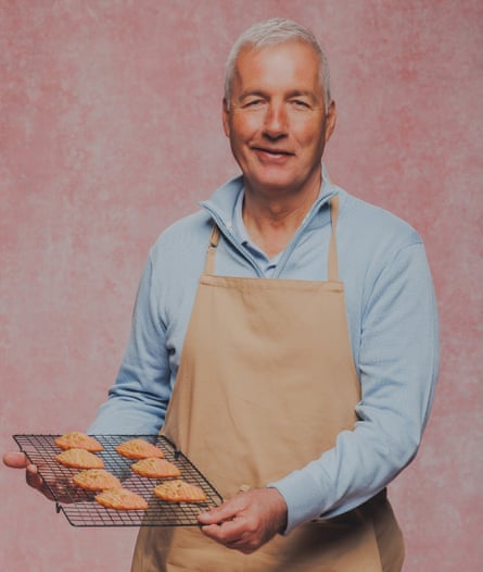 An older man with grey hair holds up a cooling rack with cookies on it.
