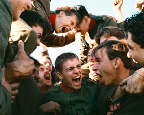 A group of young soldiers huddle together, looking down at a central figure who laughs with his mouth wide open