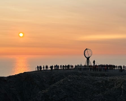 People and a globe-like structure are silhouetted against a pale orange sky as the sun sets to the left