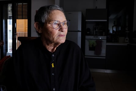 A serious-looking older white woman sits in a darkened room looking out of the window. She wears a yellow ribbon badge – a symbol of the campaign for the return of Israeli hostages