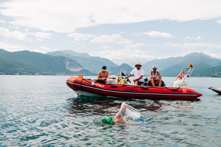 Neil Gilson swims across a lake. Three men are in an orange inflatable dinghy with outboard motor and travelling beside him. There are mountains in the background behind the lake.