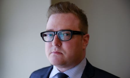 White man with short, blond coiffed hair and black-framed glasses stands against wall and looks very seriously at the camera.