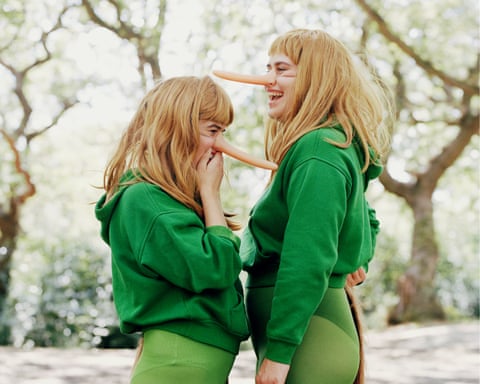 Two women in green hoodies and tights wear fake long noses and tails, laughing together outdoors