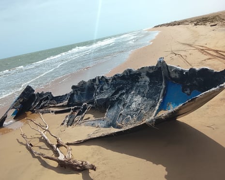 A burnt-out boat on a sandy beach feet from the ocean.