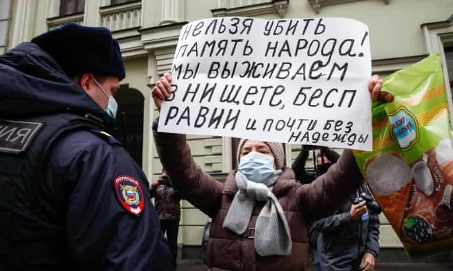 Masked woman holding sign reading: ‘It is impossible to murder the memory of a people. We are surviving in poverty, powerlessness and almost without hope.’