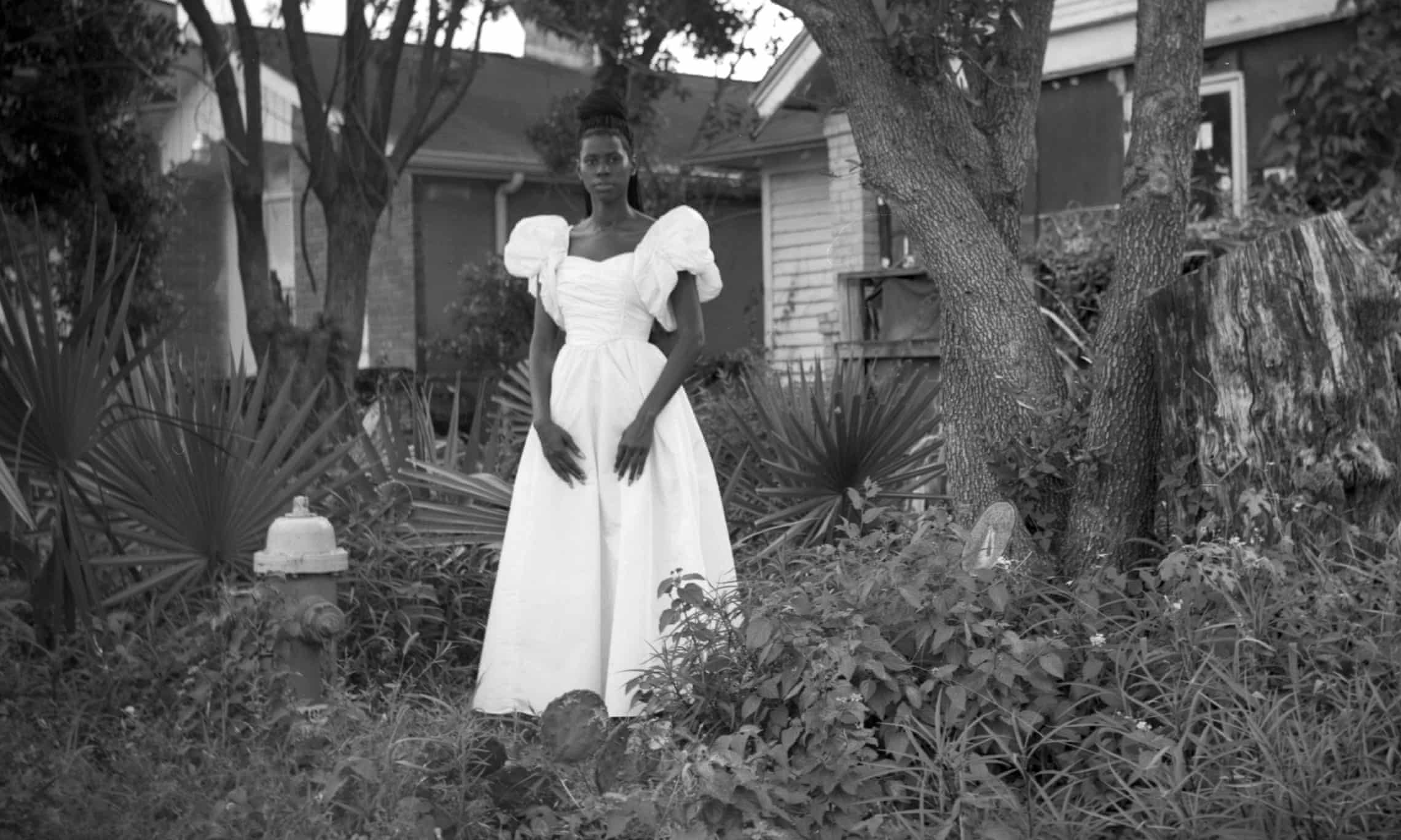 A black and white photo in which a young black woman, her hair piled up and dressed in a long white dress, gazes directly at the camera against a background of a clapboard house, palm trees, and - oddly - a fire hydrant