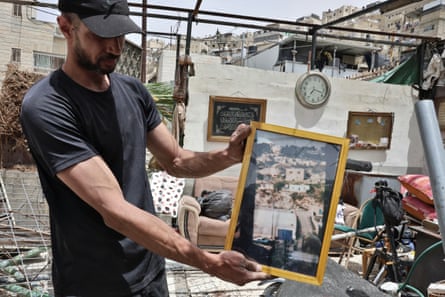 Jalal al-Tawil shows a photo of his home as he stands in the rubble of a room. One wall still stands in the background with a framed Islamic script and a clock.