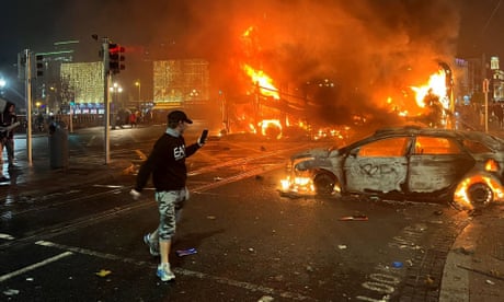 A young man crissing rthe street films the burning wreckage of a car and bus.