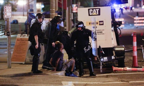a group of police officers in riot gear surround a man sitting on a curb in handcuffs