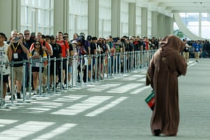 A person wearing a long brown hooded cloak walks as people in line look on