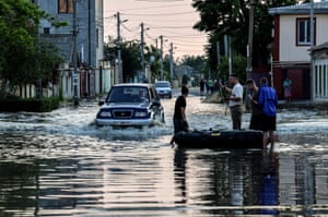 People stand next to an inflatable boat as a car drives past them in a flooded street. Ukraine has accused Russian forces of destroying the critical dam and hydroelectric power plant on the Dnipro River.