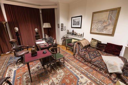 view of living room showing desk, leather chairs, paintings, a patterned rug, shelves with artefacts including busts and figurines, and a couch covered with rugs and embroidered cushions.