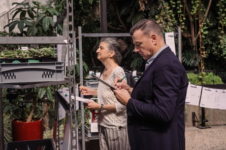 A middleaged woman and man stand in front of tiered shelves of plants with clipboards, filling in their forms. The man wears a dark suit and looks as if he is seriously thinking.