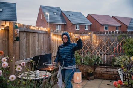 A wide-view shot of a woman grilling food on a barbecue, she is standing in a garden in the rain, she is holding serving tongs in one hand and a glass of beer in the other.