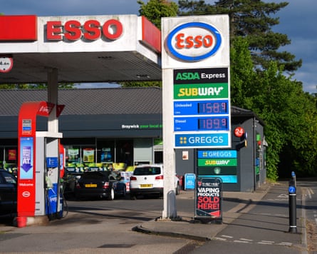 An Esso petrol station with fuel price display showing unleaded and diesel prices, with Greggs and Subway signs visible.