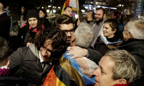 A rowd of people embrace each other and smile, with rainbow flags being worn and waved.