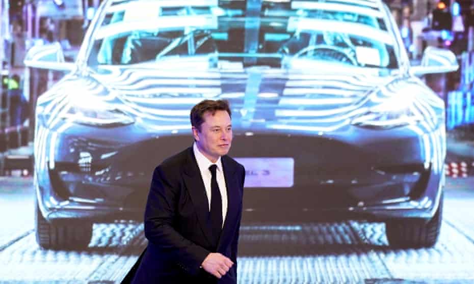 Elon Musk walks next to a screen showing an image of Tesla Model 3 car