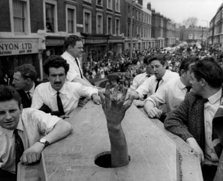 A hand emerging from a hole in a coffin being carried through a street full of people watching on.