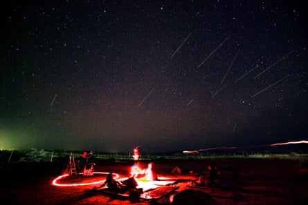 People sitting in an empty landscape as time-lapse photography captures shooting stars in the night sky 