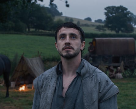 Paul Mescal standing in a field with a tent and firepit and other items behind him