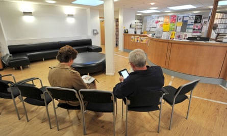 Patients sitting in the waiting room at a GP practice, one reading, the other on their phone
