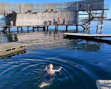 A woman swimming in a sea pool with others on wooden platforms beyond.