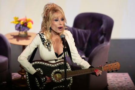 Dolly Parton singing into a microphone and playing guitar, successful beforehand of a mini array and 2 armchairs