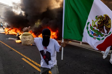 A demonstrator holds a soft mask that looks like Donald Trump and a Mexican flag in front of burning Waymo cars as protesters clash with law enforcement near the federal building during a protest in response to federal immigration operations in Los Angeles, California, on 8 June, 2025.