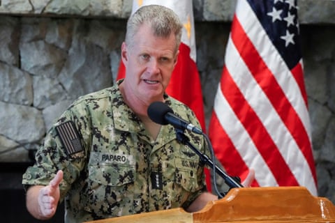 a man in military fatigues speaking in front of a US flag