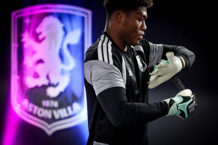 Aston Villa goalkeeper Zion Suzuki adjusts his gloves beside an illuminated club crest