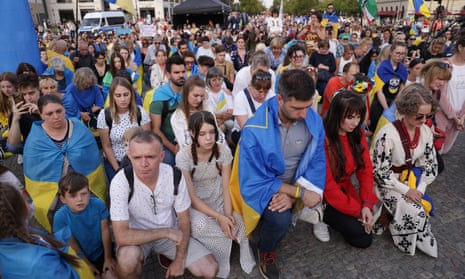 A crowd of people, many of whom are draped in the Ukrainian flag, kneel on the ground