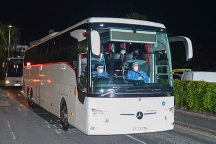A white coach pulls up in darkness at the hospital. Its driver and other people in the front can be seen wearing face masks and blue coveralls.