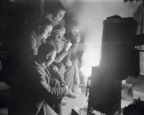 A group of people in winter clothing gather around a wood-burning stove, its fire illuminating their faces