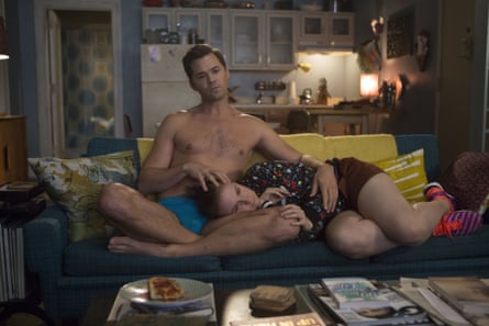 A topless man consoles a woman with her head in his lap while sitting on a sofa