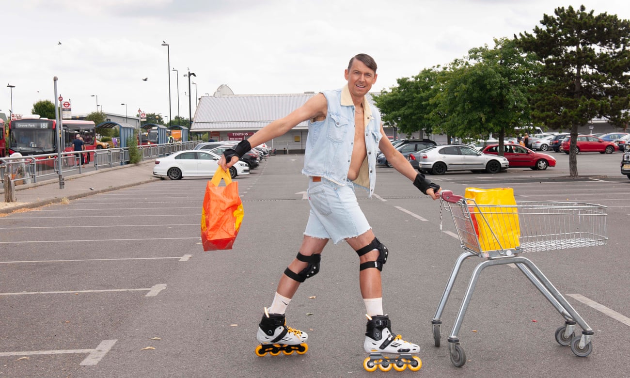 Tim Jonze on Rollerblades and wearing a cut-off demin jacket and jean shorts, pushing a trolley in a supermarket car park