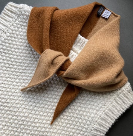 A camel coloured wool scarf is placed on a beige knitted vest and set on a dark grey background