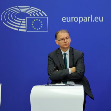 Philippe Lamberts standing with his arms folded behind a podium, against a bright blue wall with the EU flag on it and the text "europaul.eu"