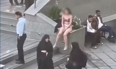 A woman in her underwear sits on a wall while others, including veiled women, walk past
