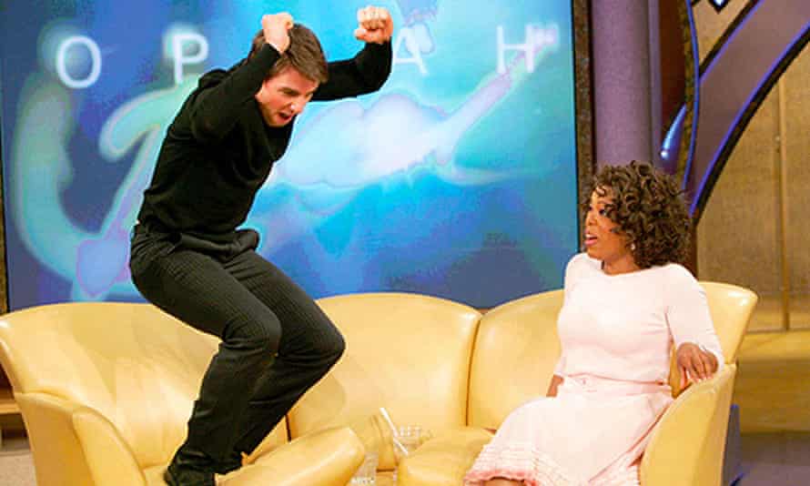 Tom Cruise standing with his arms raised at one end of a three-seat sofa, with Winfrey looking shocked at the other end.