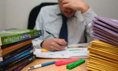A harassed-looking schoolteacher marks work next to piles of classroom books
