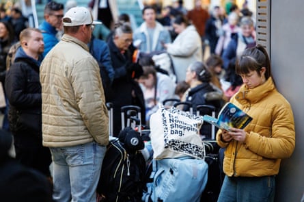 A woman reads a book while leaning against a pillar, as other people stand around with luggage