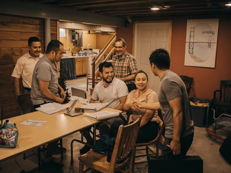 A group of four Latino men and one woman congregate around a dining table in a house. One white man, pastor Dan Krahenbuhl, stands in the background.