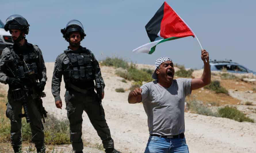 A Palestinian shouts while waving a flag as soldiers look on