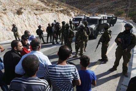 Palestinian men stand in front of Israeli soldiers and vehicles blocking a road