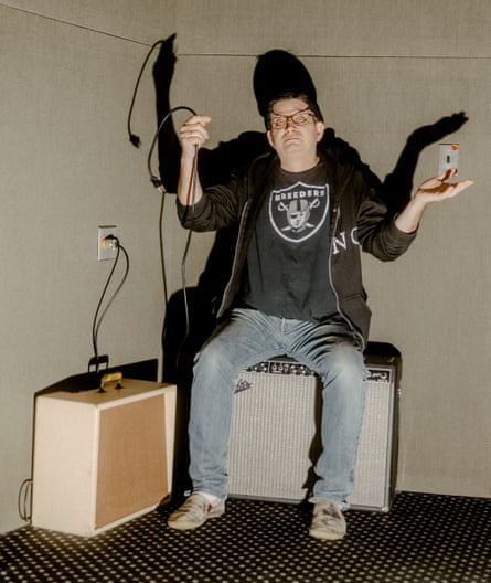 Steve Albini, wearing glasses, a T-shirt and a hoody, sitting on a loudspeaker and holding up a cable, looking bemused 
